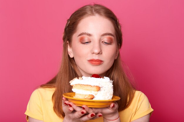 junges Mädchen, schaut auf Teller mit Stück Geburtstagstorte isoliert auf rosa, will leckeres Dessert essen, trägt gelbes T-Shirt, hat perfekte Frisur, posiert.