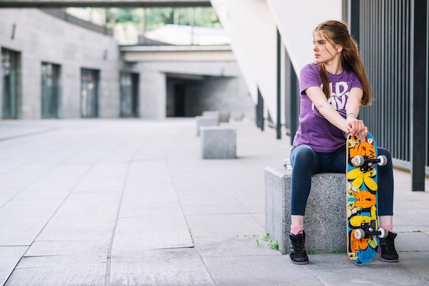 Junges Mädchen sitzt mit einem bunten Skateboard, das nach rechts schaut