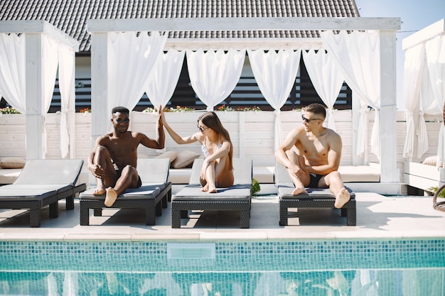 Junges Mädchen und zwei ihrer gemischtrassigen männlichen Freunde, die sich auf Sonnenliegen in der Nähe des Swimmingpools entspannen?