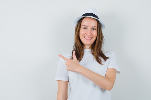 Junges Mädchen zeigt auf die obere linke Ecke im weißen T-Shirt, im Hut und schaut lustig. Vorderansicht.