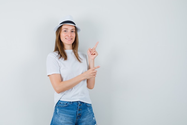 Junges Mädchen zeigt auf die obere rechte Ecke in weißem T-Shirt, Hut, Shorts und schaut lustig. Vorderansicht.