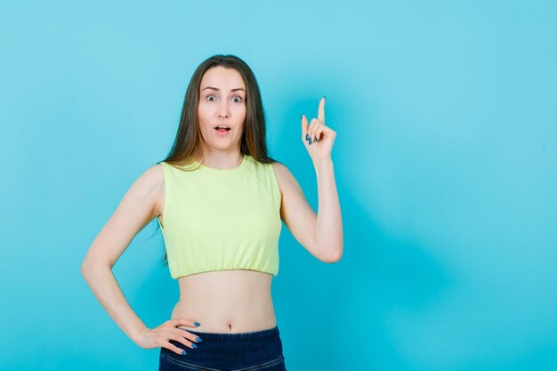 Junges Mädchen zeigt mit dem Zeigefinger nach oben und legt die andere Hand auf die Taille auf blauem Hintergrund