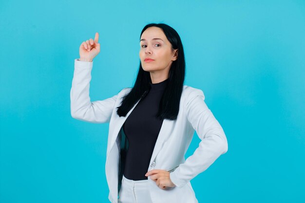 Junges Mädchen zeigt mit dem Zeigefinger nach oben und legt die Hand auf die Taille auf blauem Hintergrund