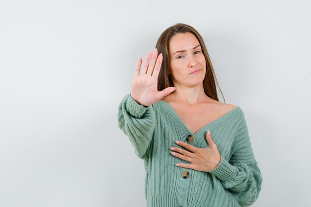 Junges Mädchen zeigt Stoppschild, hält eine Hand über Brust in Strickwaren und schaut charmant, Vorderansicht.