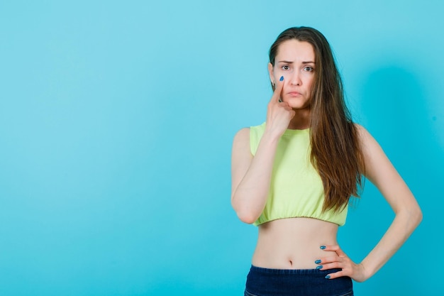 Junges Mädchen zeigt traurige Mimikry, indem es den Zeigefinger auf die Wange auf blauem Hintergrund hält