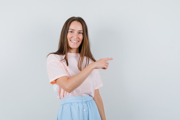 Junges Mädchen zeigt zur Seite in T-Shirt, Rock und schaut fröhlich. Vorderansicht.