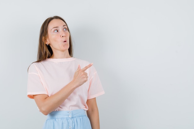 Junges Mädchen zeigt zur Seite in T-Shirt, Rock und schaut überrascht, Vorderansicht.