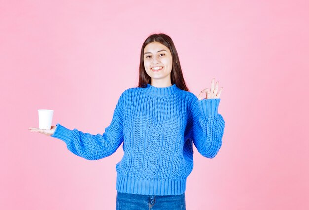 junges mädchenmodell mit einem plastikbecher, der eine ok geste an der rosa wand zeigt.