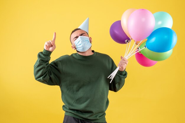 Junges Männchen der Vorderansicht, das bunte Luftballons in Maske auf gelbem Hintergrund hält