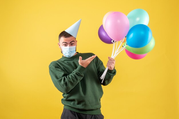 Junges Männchen der Vorderansicht, das bunte Luftballons in Maske auf gelbem Hintergrund hält