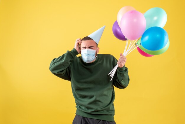 Junges Männchen der Vorderansicht, das bunte Luftballons in Maske auf gelbem Hintergrund hält