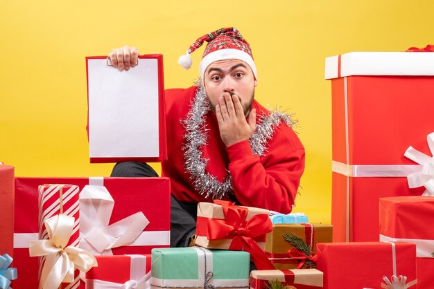 Junges Männchen der Vorderansicht, das um Weihnachtsgeschenke mit Hinweis auf gelbem Hintergrund sitzt