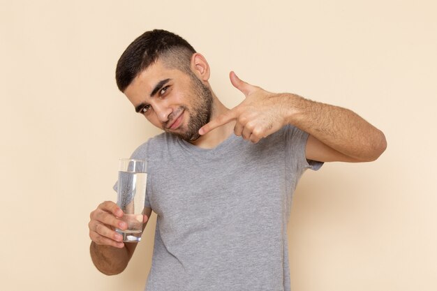 Junges Männchen der Vorderansicht im grauen T-Shirt, das Glas Wasser auf Beige hält