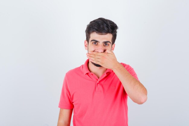 Junges Männchen im rosa T-Shirt, das Hand auf den Mund hält und fröhlich aussieht, Vorderansicht.