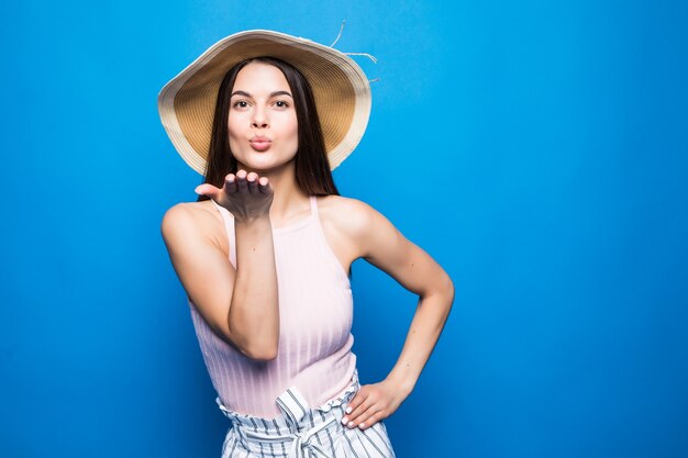 Junges modisches attraktives Mädchen in Sonnenbrille und Strohhut sendet Luftkuss zur Kamera. Schöne verspielte und glückliche Frau mit roten Lippen auf blauer Wand. mit Kopierplatz.