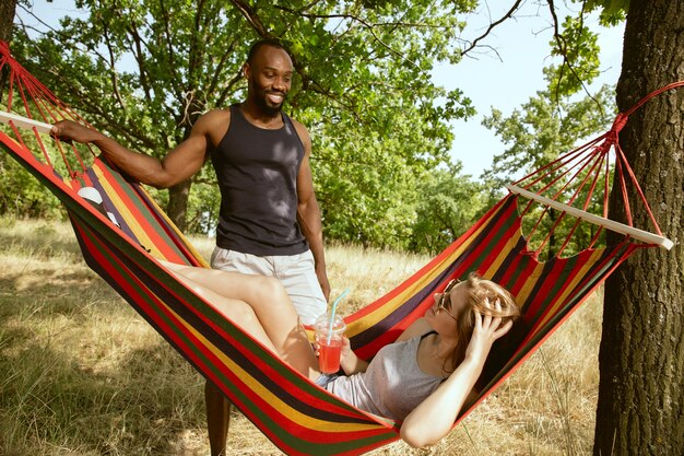 Junges multiethnisches internationales romantisches Paar draußen an der Wiese im sonnigen Sommertag. Afroamerikanermann und kaukasische Frau, die zusammen Picknick haben. Konzept der Beziehung, Sommerzeit.