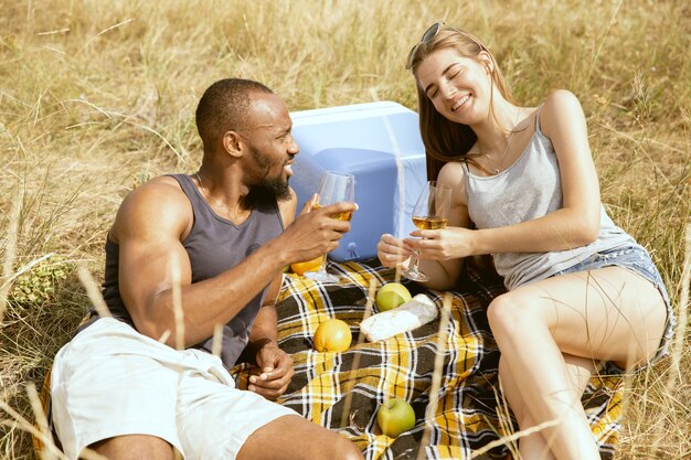 Junges multiethnisches internationales romantisches Paar draußen an der Wiese im sonnigen Sommertag. Afroamerikanermann und kaukasische Frau, die zusammen Picknick haben. Konzept der Beziehung, Sommerzeit.