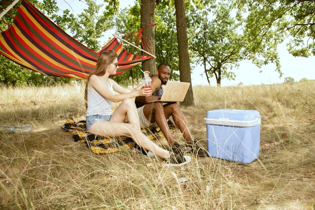 Junges multiethnisches internationales romantisches Paar draußen an der Wiese im sonnigen Sommertag. Afroamerikanermann und kaukasische Frau, die zusammen Picknick haben. Konzept der Beziehung, Sommerzeit.