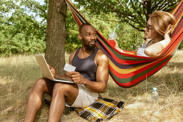 Kostenloses Foto junges multiethnisches internationales romantisches paar draußen an der wiese im sonnigen sommertag. afroamerikanermann und kaukasische frau, die zusammen picknick haben. konzept der beziehung, sommerzeit.