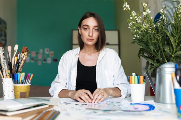 Junges nachdenkliches Mädchen mit dunklen Haaren sitzt am Schreibtisch mit Bildern, die verträumt in die Kamera schauen, während sie zu Hause zeichnen