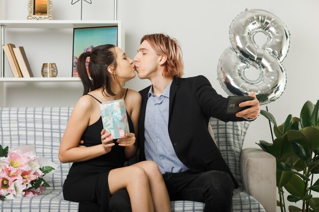 Junges Paar am glücklichen Frauentag, der Geschenk hält, macht ein Selfie, das auf dem Sofa im Wohnzimmer sitzt