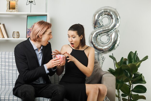 Junges Paar am glücklichen Frauentag lächelnder Kerl gibt einer aufgeregten Frau, die auf dem Sofa im Wohnzimmer sitzt, ein Geschenk