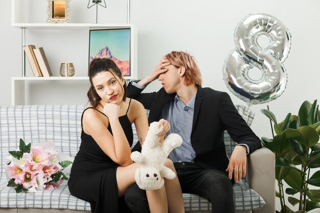 Junges Paar am glücklichen Frauentag mit Teddybär bedecktes Gesicht mit der Hand, die auf Sofa im Wohnzimmer sitzt