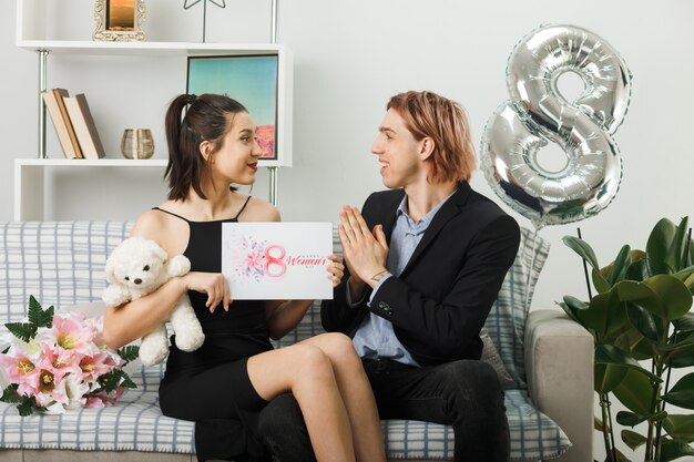 Junges Paar am glücklichen Frauentag mit Teddybär und Postkarte auf dem Sofa im Wohnzimmer sitzend