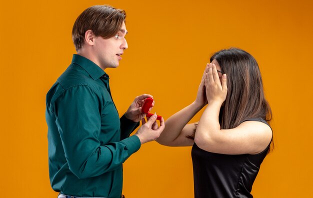 Junges Paar am Valentinstag beeindruckte den Mann, der den Augen des Mädchens mit der Hand isoliert auf orangefarbenem Hintergrund Ehering gab