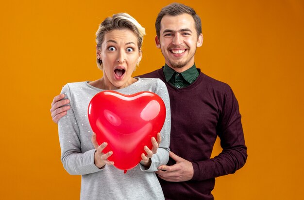 Junges Paar am Valentinstag, der hinter überraschtem Mädchen mit Ballon auf orangefarbenem Hintergrund steht
