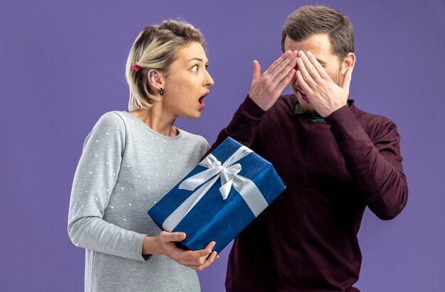 Junges Paar am Valentinstag erschrockenes Mädchen, das dem Kerl eine Geschenkbox gibt, die auf blauem Hintergrund isoliert ist