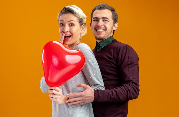 Junges Paar am Valentinstag lächelnder Kerl umarmte lachendes Mädchen mit Herzballon isoliert auf orangem Hintergrund