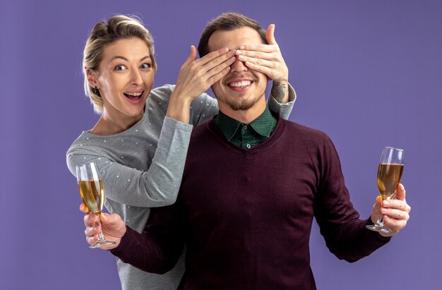 Junges Paar am Valentinstag - lächelndes Mädchen bedeckte Kerl Etes mit Gläsern Champagner isoliert auf blauem Hintergrund