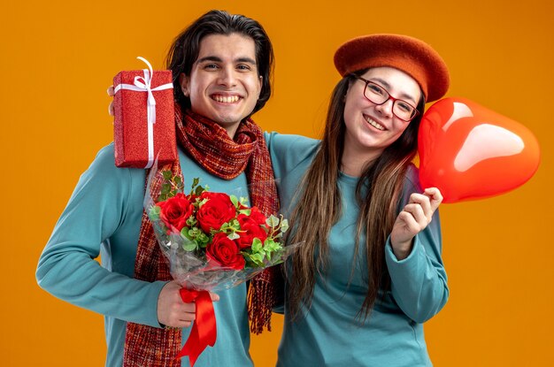 Junges Paar am Valentinstag lächelndes Mädchen mit Herzballon umarmte Kerl mit Blumenstrauß isoliert auf orangem Hintergrund