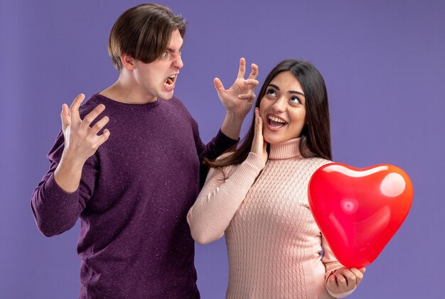 Junges Paar am Valentinstag wütender Kerl, der aufgeregtes Mädchen mit Herzballon anschaut, der Hand auf die Wange legt, isoliert auf blauem Hintergrund