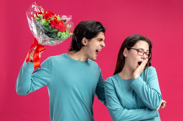 Junges Paar am Valentinstag wütender Kerl, der Blumenstrauß anhebt, bedeckt den Mund mit der Hand, die auf rosa Hintergrund isoliert ist isolated