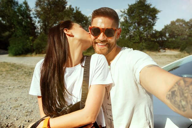 Junges Paar, das an sonnigem Sommertag auf dem Auto Urlaub macht. Frau und Mann machen Selfie im Wald und sehen glücklich aus. Konzept der Beziehung, Urlaub, Sommer, Urlaub, Wochenende, Flitterwochen.