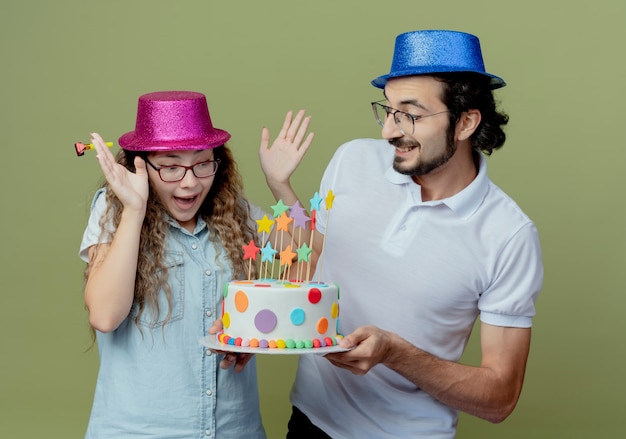 Junges Paar, das den lächelnden Kerl des rosa und blauen Hutes trägt, gibt dem überraschten Mädchen, das auf Olivgrün isoliert wird, Geburtstagstorte
