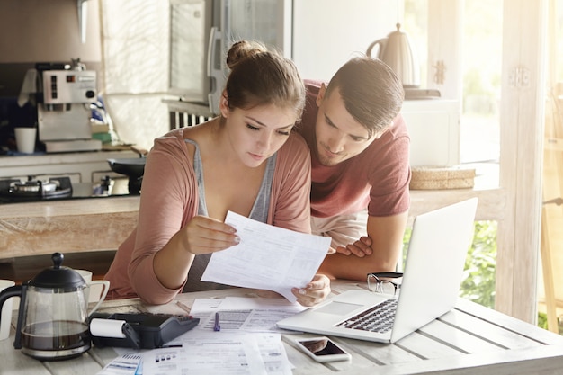 Junges Paar, das Finanzen verwaltet und ihre Bankkonten mit Laptop und Taschenrechner in der modernen Küche überprüft. Frau und Mann machen Papierkram zusammen