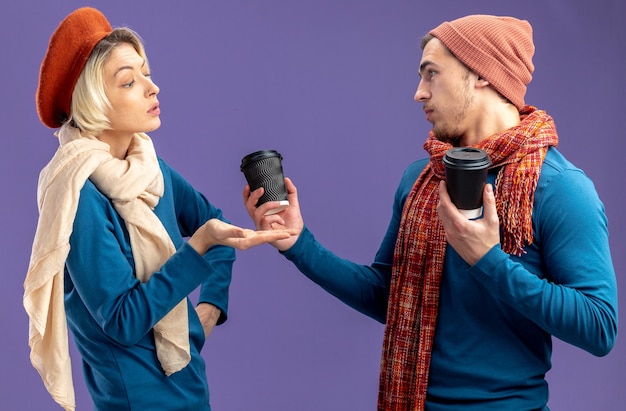 Junges Paar, das Hut mit Schal am Valentinstag trägt, beeindruckte Mädchen, die mit der Hand auf einen Mann mit einer Tasse Kaffee auf blauem Hintergrund zeigen