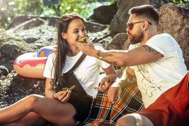 Junges Paar, das Picknick am Flussufer im sonnigen Tag hat. Frau und Mann verbringen Zeit auf der Natur zusammen. Spaß haben, essen, spielen und lachen. Konzept der Beziehung, Liebe, Sommer, Wochenende.