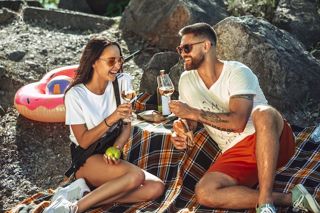 Junges Paar, das Picknick am Flussufer im sonnigen Tag hat. Frau und Mann verbringen Zeit auf der Natur zusammen. Spaß haben, essen, spielen und lachen. Konzept der Beziehung, Liebe, Sommer, Wochenende.