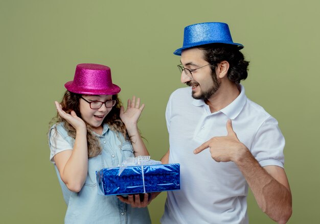 Junges Paar, das rosa und blauen Hut lächelnden Kerl trägt, gibt Geschenkbox zu überraschtem Mädchen lokalisiert auf Olivgrün