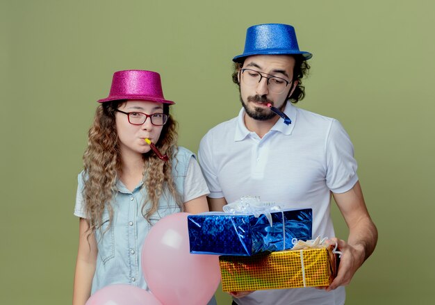 Junges Paar, das rosa und blauen Hut trägt, der Pfeife und Kerl bläst und Geschenkboxen lokalisiert auf olivgrüner Wand hält