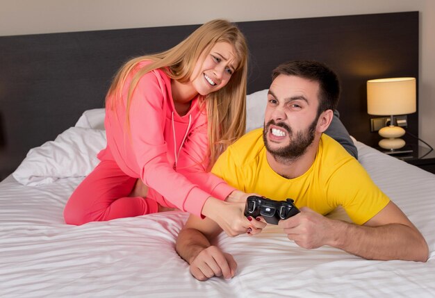 Junges Paar, das Videospiele im Bett spielt. Guy spielt ein Videospiel, sie beschwert sich, dass sie nicht aufgepasst hat