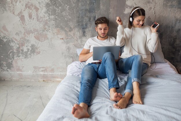 Junges Paar, das zu Hause im lässigen Outfit auf Bett sitzt, beschäftigter Mann, der freiberuflich auf Laptop arbeitet, Frau, die Musik auf Kopfhörern hört, Zeit zusammen verbringend