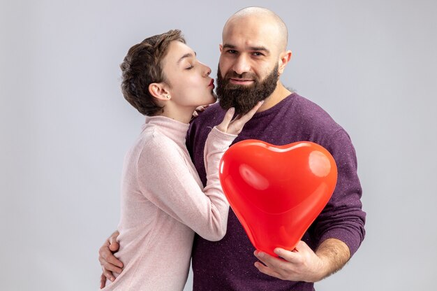 junges Paar in der Freizeitkleidung glückliche Frau, die ihren bärtigen Freund mit Herzformballon küsst, der Valentinstag feiert, der über weißem Hintergrund steht