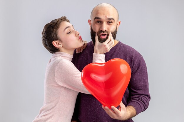 junges Paar in der Freizeitkleidung glückliche Frau, die Wangen ihres schüchternen lächelnden bärtigen Freundes berührt, der herzförmigen Ballon hält, der Valentinstag feiert, der über weißer Wand steht