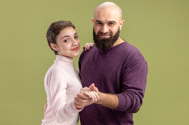 junges Paar in Freizeitkleidung Frau mit kurzen Haaren und bärtigem Mann glücklich in der Liebe tanzen Valentinstag feiern über grüne Wand stehen