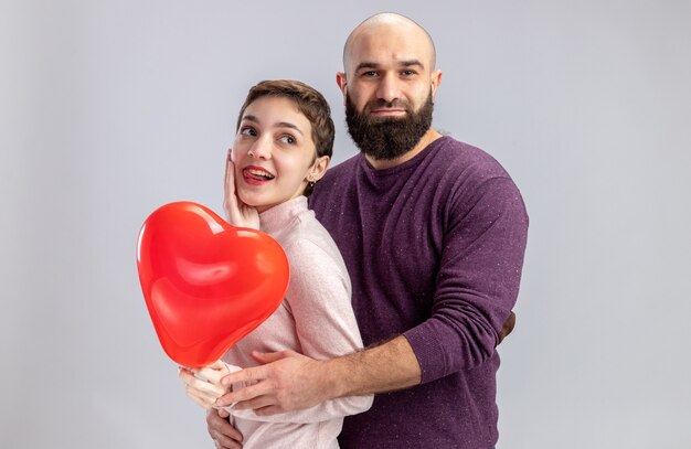 junges Paar in lässiger Kleidung Mann und Frau, die herzförmigen Ballon glücklich in Liebesmeilen halten, der fröhlich Valentinstag feiert, der über weißer Wand steht